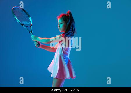 Ein kleines Mädchen, Anfänger Tennisspielerin posiert isoliert auf blauem Studiohintergrund in Neonlicht. Sport, Studium, Kinderkonzept Stockfoto