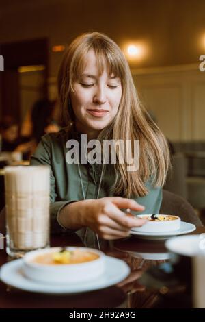 Lächelnde Frau, die im Café Creme Brulee isst. Blick auf das Dessert. Gemütliche Atmosphäre Stockfoto