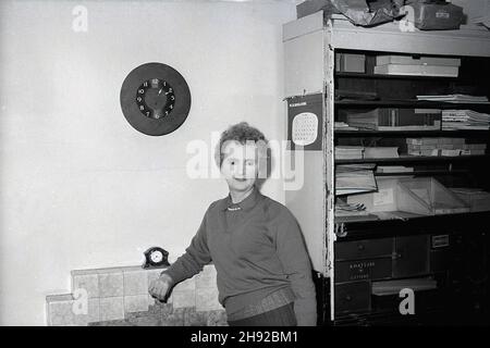 1959, historisch, stehend an einem gefliesten Kamin, eine Angestellte in der Verwaltungsabteilung von Mullards Limited, einem führenden britischen Elektronikhersteller der Ära. Ursprünglich von Captain Stanley R. Mullard im Jahr 1920 gegründet, wurden in einer Partnerschaft mit dem niederländischen Elektronikunternehmen Phillips im Jahr 1923 Funkwerte unter der Marke Phillips-Mullard ermittelt. Phillips übernahm 1927 die Kontrolle über die Partnerschaft. Zu einem Zeitpunkt im Jahr 1960s war die Mullard-Fabrik in Blackburn, Lancashire, die größte in Europa, die Werte für Radio und Fernsehen produzierte. Der alte Aktenschrank auf der rechten Seite hat einen Kasten mit der Aufschrift „Amateur Letters“. Stockfoto