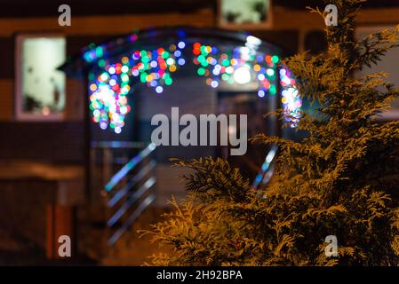 Bunte Girlande in verschwommener Form am Eingang eines Mehrfamilienhauses. Thuja am Abend vor dem Eingang, für den dekoriert ist Stockfoto