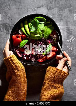 Frischer Salat aus violettem Kohl, Tomaten und Avocado auf einem Teller und weibliche Hände in einem Strickpullover Stockfoto