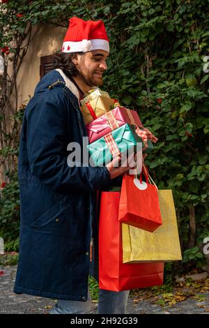 Weihnachtseinkäufe. Der junge Mann im Weihnachtsmannhut hat einen besorgten Ausdruck, als er seine neu gekauften Geschenke bringt, er hat Angst, t fallen zu lassen Stockfoto