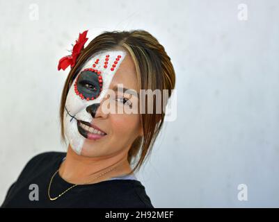 Die junge attraktive mexikanische Frau malt am Tag der Toten die rechte Hälfte ihres Gesichts mit einem schaurigen weißen Catrina Face Make-up und schaut den Betrachter an. Stockfoto