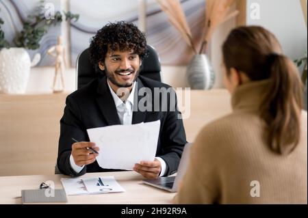 Selbstbewusst erfolgreiche indische männliche HR-Manager, Unternehmer, führt Interview Mädchen in einem modernen Büro, trägt formelle Kleidung, suchen und hören einen Job-Kandidaten, hält einen Lebenslauf in den Händen Stockfoto