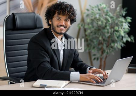 Porträt eines positiven charismatischen jungen indischen Geschäftsmannes mit lockigen Haaren, der in Geschäftskleidung an seinem Schreibtisch im Büro sitzt und mit einem freundlichen Lächeln auf die Kamera blickt Stockfoto