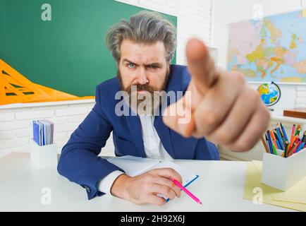 Wütender männlicher Lehrer im Klassenzimmer zeigt mit dem Finger. Ernsthafter bärtiger Mann in Anzug, der im Unterricht am Tisch sitzt. Stockfoto