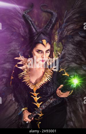 MALEFICENT COSPLAY. PORTRÄT, DUNKLER CHARAKTER MIT SCHARFEN HÖRNERN UND STARKEN FLÜGELN. BÖSE FEE IN SCHWARZEM KLEID. GELBE AUGEN. VERTIKAL. LILA FARBE. Stockfoto