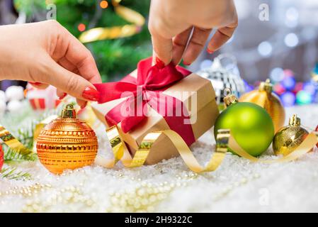 Geschenkverpackung mit einer Schleife in den Händen eines Mädchens mit einer Serpentine und Weihnachtsspielzeug auf dem Hintergrund eines Weihnachtsbaums und Bokeh für das neue Jahr Stockfoto