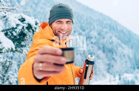 Ein Mann mittleren Alters bietet einen heißen Drink Tasse von einem Thermoskanne gekleidet in einem hellen orange Softshell Jacke während Er Trekking die Winterberge rou Stockfoto
