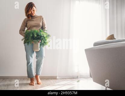 Eine junge Frau zog einen warmen, gemütlichen Pullover an, der die Tonvase voller frisch geschnittener, grüner Tannenbaum-Nadelzweige in der Nähe einer weißen Wand im Heim-Liv hält Stockfoto