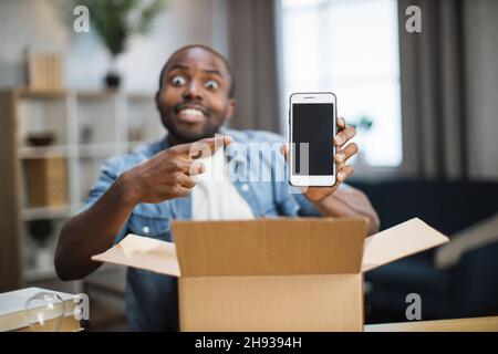 afroamerikanischer Blogger, der auf die Kamera schaut und mit dem Finger auf das neue moderne Smartphone zeigt. Fröhlicher junger Mann, der mit offenem Papierkarton am Schreibtisch saß. Stockfoto