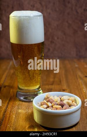 Ein Glas Lagerbier mit Erdnüssen auf dem Tisch und in einer weißen Schüssel auf einem Holztisch. Stockfoto