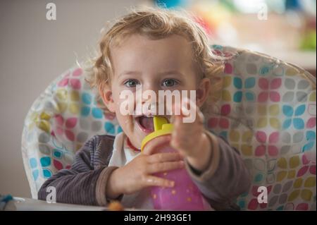 Ein 2-jähriges Mädchen lächelt, während sie auf einem Hochstuhl sitzt und mit einer Babyflasche spielt. Stockfoto