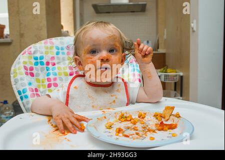 Ein kleines Mädchen (15 Monate alt) isst eine Mahlzeit aus süßem Mais und Tomatensauce auf unordentliche Weise und spielt mit ihrem Essen auf einem Hochstuhl in ihrem Haus. Stockfoto