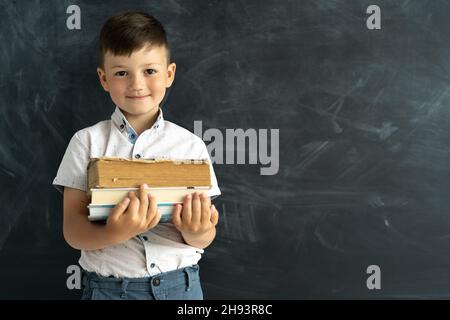 Zurück zur Schule. Schuljunge, der mit Büchern auf dem Hintergrund einer schwarzen Kreidetafel mit Platz für Text steht. Fakultät Student smart child in der Stockfoto