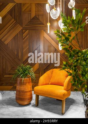 Moderner orangefarbener Sessel, hoher Holztopfern und moderner Kronleuchter aus Glas in einem Saal mit dekorierter Holzverkleidung und weißem Marmorboden, der von grünen Büschen eingerahmt ist Stockfoto