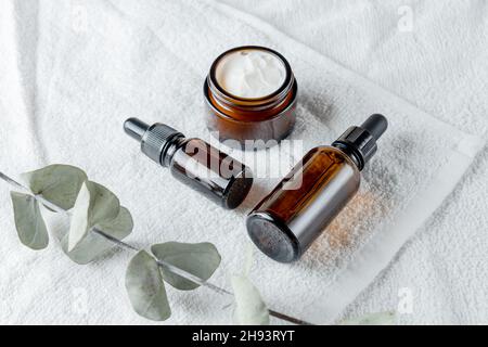 Braune Glasflaschen und Eukalyptuszweig auf weißem Handtuchhintergrund. Umweltfreundliche Bio-Naturkosmetik für Haut- und Körperpflege. Speicherplatz Kopieren. Stockfoto