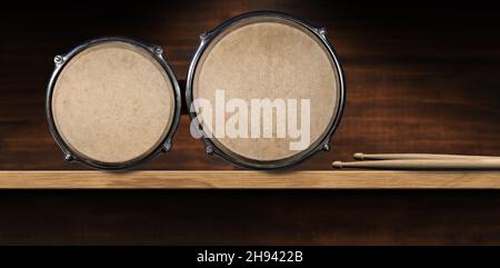 Nahaufnahme von Bongo-Trommeln und einem Paar Drumsticks, über einem Holzregal mit Platz für Kopien. Schlaginstrument. Fotografie. Stockfoto