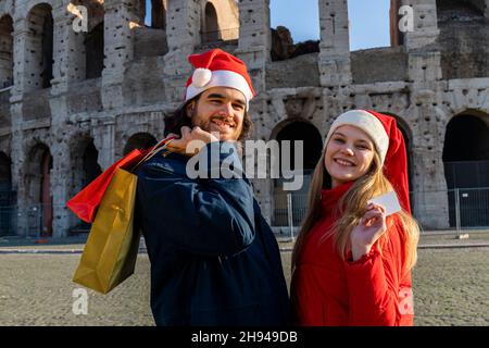 Weihnachtseinkäufe. Das lächelnde junge Paar hat gerade Weihnachtsgeschenke gekauft. Er bringt die Umschläge mit Geschenken und sie zeigt ihre Kreditkarte Stockfoto