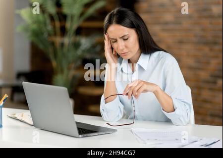 Erschöpfte kaukasische Frau, Managerin, Sekretärin, müde von der Arbeit mit dem Laptop, während sie an ihrem Arbeitsplatz sitzt. Überarbeitete Geschäftsfrau reibt sich den Nasenrücken, hält eine Brille in der Hand, hat Migräne Stockfoto