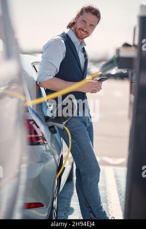 Der junge Geschäftsmann wartet darauf, dass sein Elektroauto an der Ladestation geladen wird Stockfoto