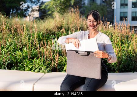Lächelnde Geschäftsfrau, die ihren Laptop aus der Aktentasche nimmt, die in einem Park sitzt, Konzept des digitalen Unternehmers und des urbanen Lebensstils, Copy Spa Stockfoto