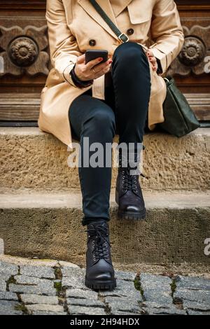 Frau, die in der Stadt auf Treppen sitzt und Smartphone benutzt. Stilvolle Frau in Trenchcoat, schwarzer Jeans und Lederstiefeln. Straßenstil Stockfoto