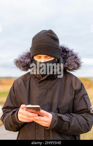 Ein junger Mann, der während der Pandemie des Corona-Virus Covid-19 eine schwarze, waschbare Gesichtsmask trug, während er sein Mobiltelefon benutzte Stockfoto