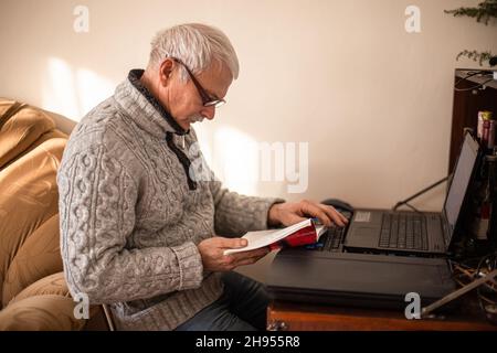 Der weißhaarige ältere Mann liest ein Buch, das neben einem Tisch mit einem Laptop sitzt. Der alte Mann liest zu Hause die Anweisungen für ein modernes Gadget. Stockfoto