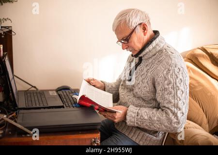 Der weißhaarige ältere Mann liest ein Buch, das neben einem Tisch mit einem Laptop sitzt. Der alte Mann liest zu Hause die Anweisungen für ein modernes Gadget. Stockfoto