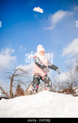 Kind wirft Schneebälle im Winterpark. Kleines Mädchen im rosa Winteranzug spielt und reitet auf der Schneerutsche gegen den blauen klaren Himmel am sonnigen Wintermorgen Stockfoto