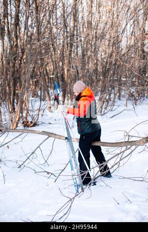 Junge Frau macht Sport im Wald. Brunette im Winter Trainingsanzug geht entlang verschneiten Straße und hält Skistöcke und Skier in ihren Händen, Seitenansicht Stockfoto