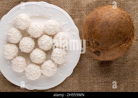 Eine natürliche Kokosnuss mit Süßigkeiten mit Kokosflocken auf einem weißen Teller auf einem Jute-Stoff, Nahaufnahme, Draufsicht. Stockfoto