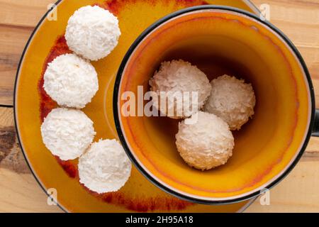 Mehrere leckere Süßigkeiten mit Kokosflocken mit einer keramischen Tasse und Untertasse auf einem Holztisch, Nahaufnahme, Draufsicht. Stockfoto