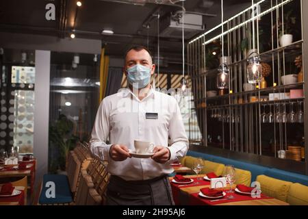 Professioneller Kellner, der während einer Coronavirus-Pandemie im Restaurant arbeitet und eine medizinische Gesichtsmaske trägt Stockfoto