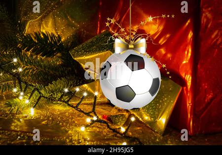 Fußball-weihnachtskugel, die vor einem beleuchteten goldenen und roten Geschenkhintergrund hängt. Weihnachtskarte für Grüße Stockfoto