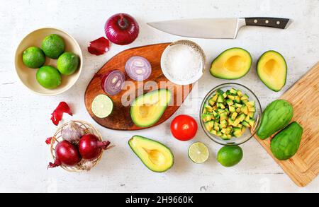 Avocado-Hälften, Stücke, Tomaten, Limetten und Zwiebeln, Tasse mit Salz - Grundzutaten der Guacamole und Kochmesser auf weißem Arbeitsbrett, flach liegend Foto Stockfoto