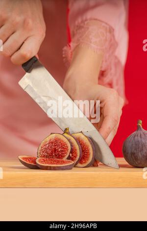 Weibliche Hände hacken frische Feigen auf einem Holzbrett auf einem Tisch mit einem großen Kochmesser auf rotem Hintergrund Stockfoto