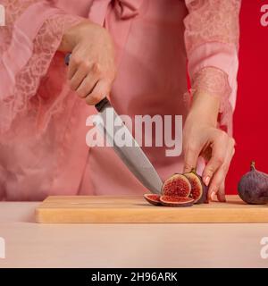 Weibliche Hände hacken frische Feigen auf einem Holzbrett auf einem Tisch mit einem großen Kochmesser auf rotem Hintergrund Stockfoto