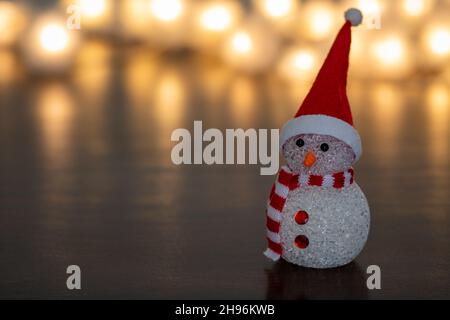 Kleine Figur eines niedlichen Weihnachtsschneemanns, der einen rot-weißen Schal und einen Weihnachtsmann-Hut mit verschwommenen Lichterketten trägt Stockfoto