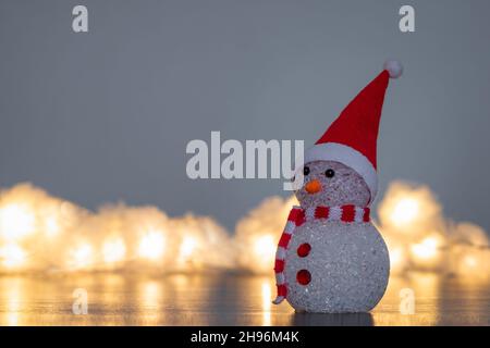 Kleine Figur eines niedlichen Weihnachtsschneemanns, der einen rot-weißen Schal und einen Weihnachtsmann-Hut mit verschwommenen Lichterketten trägt Stockfoto