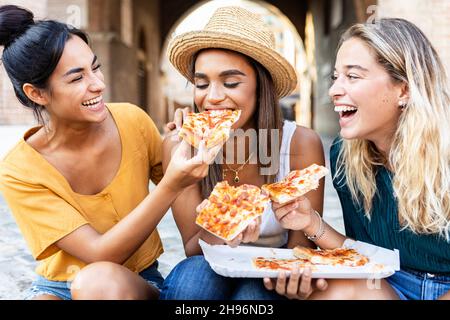 Drei fröhliche, multirassische Frauen essen Pizza auf der Straße Stockfoto