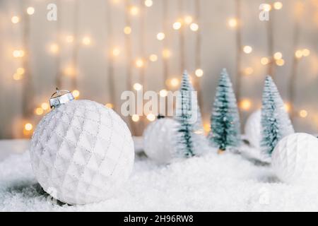 Weihnachtsmärchen Komposition mit weißen Kugeln Dekor und kleinen Weihnachtsbäumen auf dem Schnee vor warmen Lichtern Bokeh Hintergrund. Frohe Weihnachten Stockfoto