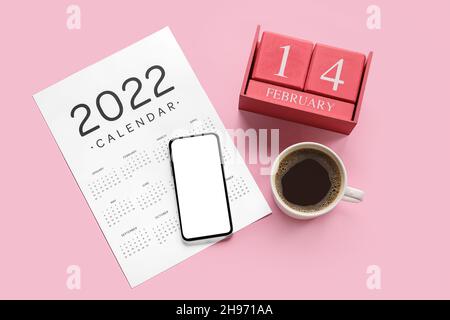 Papierkalender für 2022 Jahre und Würfelkalender mit Datum FEBRUAR 14, Mobiltelefon und Tasse Kaffee auf farbigem Hintergrund Stockfoto