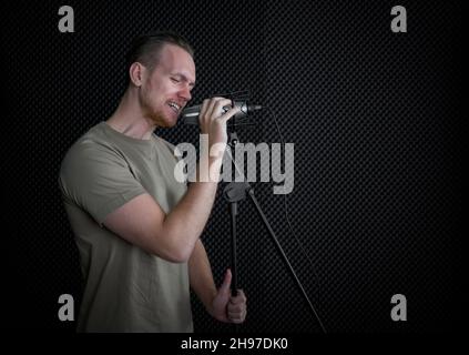 Junger Kaukasier singt vor schwarzen Schallschutzwänden. Musiker, die Musik im professionellen Aufnahmestudio produzieren. Stockfoto