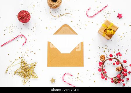 Offener brauner Umschlag mit weißer Karte und Weihnachtsdekorationen, Zuckerstöcken und Geschenkbox auf weißem Hintergrund. Draufsicht, flaches Lay, Mock Up Stockfoto