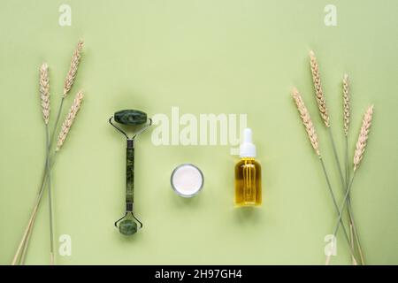 Kosmetisches Serum in Glasflasche, Cremedose, Gesichtswalze und Ähren aus Weizen auf hellgrünem Hintergrund. Hautpflege, natürliche Bio-Kosmetik-Konzept. Oben Stockfoto