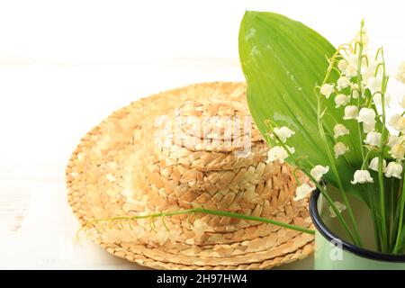 Lilien des Tals in einem emaillierten Becher mit Strohhut auf weißem Holzgrund Stockfoto