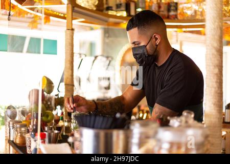 Junger lateinamerikanischer Kellner, der an der Bar arbeitet. Er trägt eine Gesichtsschutzmaske. Platz für Text. Stockfoto