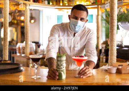 Eleganter junger lateinamerikanischer Barkeeper, der einen Cocktail an der Bar einer modernen Cocktailbar serviert. Während der Arbeit trägt er eine OP-Maske. Stockfoto